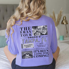 a woman sitting on a bed wearing a purple shirt