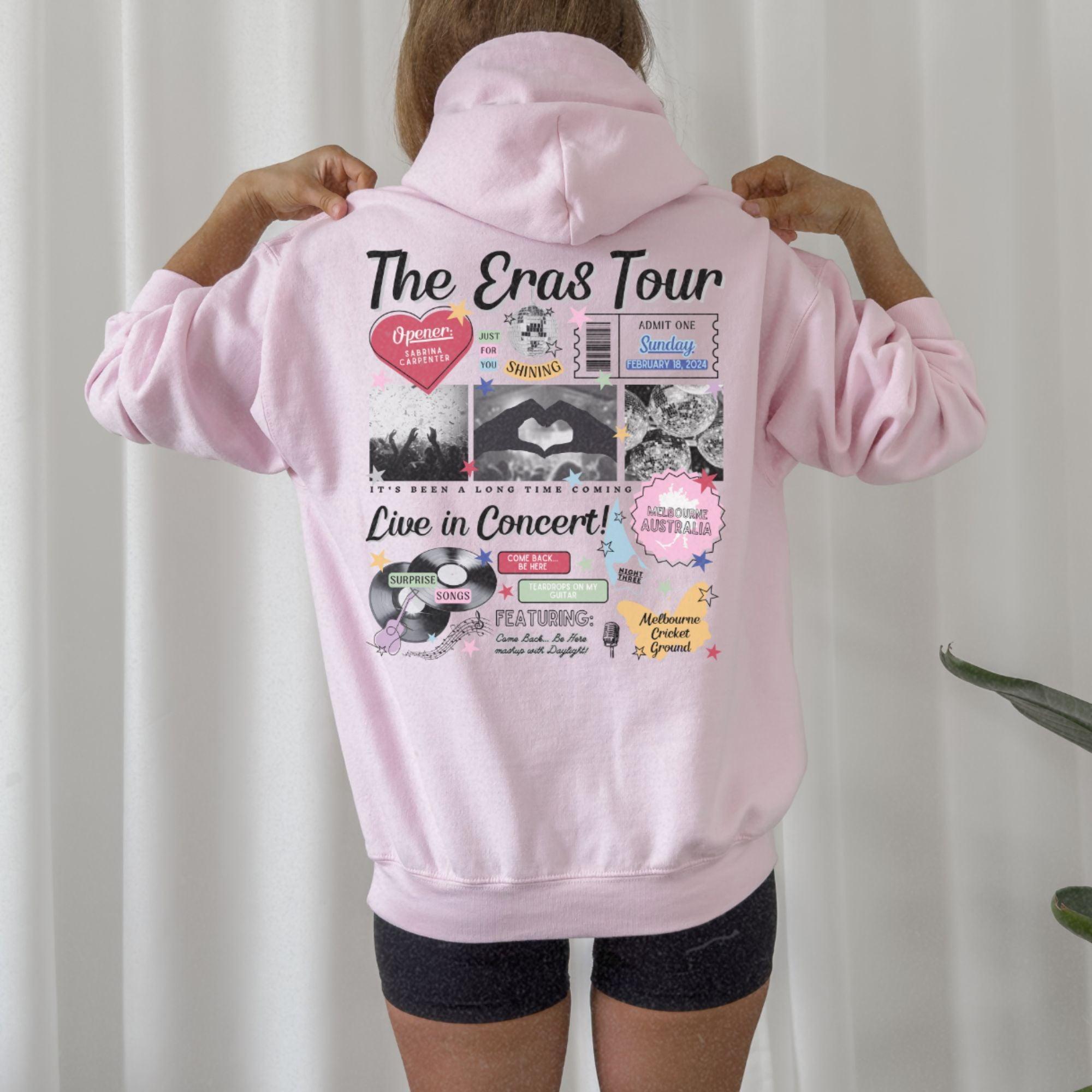 a woman wearing a pink hoodie with the words the eras tour printed on it