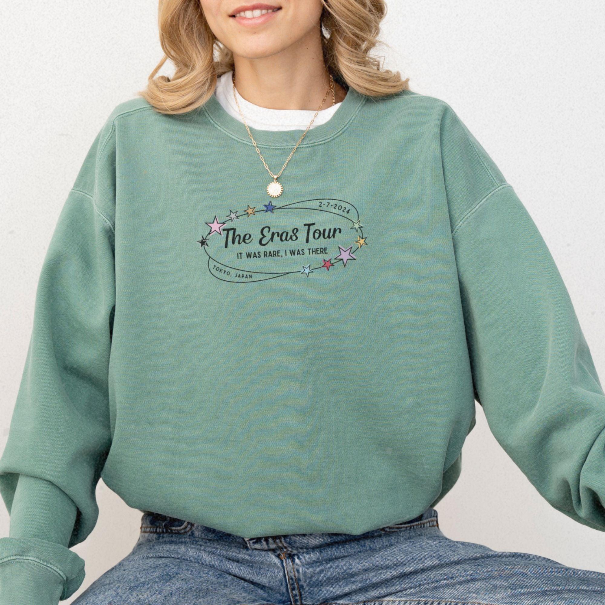 a woman wearing a green sweatshirt with the words the eras tour on it